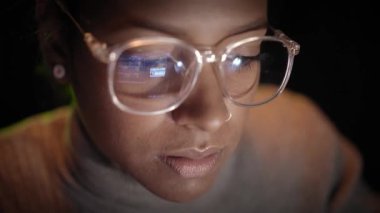 Karanlıkta dizüstü bilgisayarı üzerinde çalışan Afrikalı Amerikalı bir hacker kadının gözlüklerindeki bilgisayar ekranının yansımasına yakın çekim. Gençler ve bilgisayar ağları ve İnternet güvenliği. 