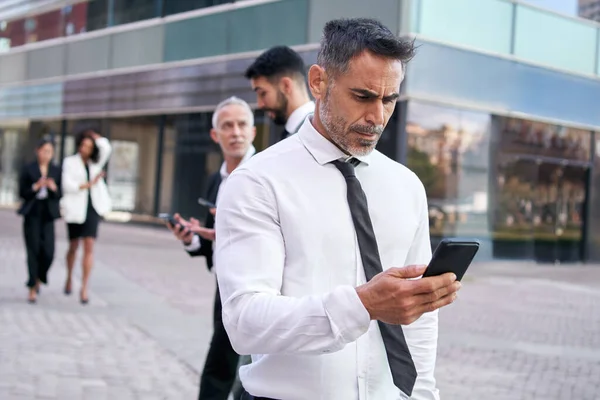 Attractive middle-aged business man looks at cell phone concentrating walking down the street in a crowd. Serious formal staff outside office center. Male professional people using mobile at work.