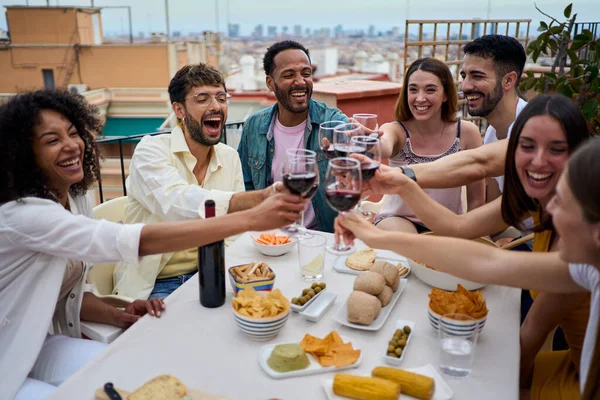 Gathering cheerful young multiracial friends toasting red wine and celebrating party on rooftop. People together drinking alcoholic beverages at snack outdoor. Millennial enjoying lunch on weekend.