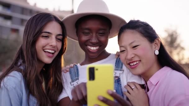 三个年轻的 面带微笑的多民族女孩在户外一起使用黄色手机 一群漂亮的朋友在暑假带着自己的手机为社交媒体服务 对装置上瘾的人 — 图库视频影像