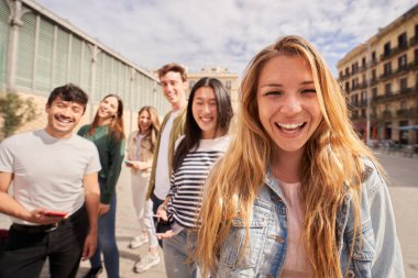 Arka plandaki sokak grubu çok ırklı arkadaşlarına bakan neşeli sarışın kadına odaklan. Birlikte gülümseyen insanlar portre yapıyor. İş arkadaşları tatilde etrafı geziyor. Z Jenerasyonu. 