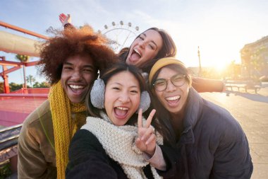 Çok kültürlü arkadaşlardan oluşan heyecanlı bir grup dışarıda birlikte kameraya bakarken komik selfie çekiyorlar. Eğlence parkında çok ırklı gençler güneşli kış tatilinde eğleniyorlar.. 