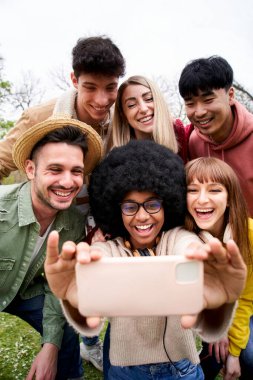Dikey gülümseyen Afrikalı afro kız selfie telefon parkı çok ırklı arkadaşlarını götürüyor. Mutlu insanlar bahar günü dışarıda arkadaşlığın tadını çıkarıyorlar. Z nesli topluluk ilişkileri, sosyal ağlar.