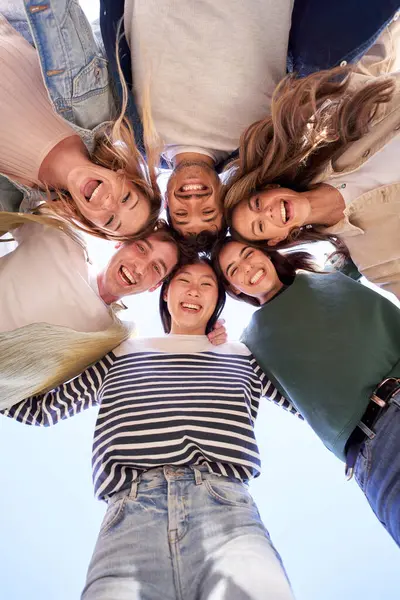 Vertical multiracial group of young people standing in circle and smiling excited at camera. Happy diverse generation z friends having fun embracing together. Low angle view sunny portrait outdoor.