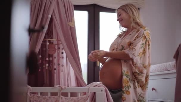 怀孕的女人把未来的婴儿衣服和爱放在一起 年轻的金发姑娘大肚子为新生儿安排房间 母亲在千禧年家庭等待新成员的出生 母亲和规划 — 图库视频影像