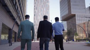 Üç takım elbiseli iş adamının şehir caddesinde yürüyüp konuşmalarını takip edin. Arkadan bakan erkek finans ortakları ofis binası dışında çokuluslu gezintiler ve iletişim. Açık hava iş ilişkileri.