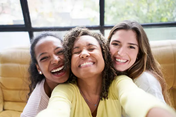 Selfie photo of three cheerful young girls posing smiling indoors. Group of attractive women together looking funny at camera for picture at home. People generation z enjoying leisure with friends.