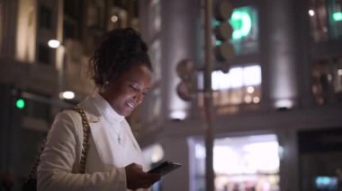 Genç Afrikalı Amerikalı kadın aydınlanmış şehir merkezinde cep telefonuna bakarak gülümsüyor. Cosmopolitan kadın ana cadde boyunca gezintiye çıkıyor. Gen z insanları cep telefonu bağımlısı.