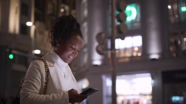 若いアフリカ系アメリカ人女性は 夜に笑顔で街の中心部を照らして携帯電話を見ています メインストリートに沿って散歩する途中のコスモポリタン女性 モバイル中毒のZ世代 — ストック動画