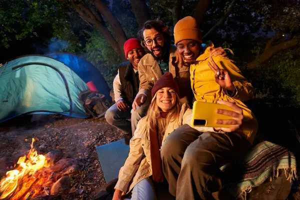 Group multi-ethnic friends smiling and taking selfie phone sitting front campfire. Young gathered together camping on winter night. Two cheerful millennial excited couples happy embracing on getaway.