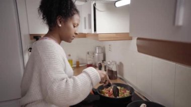 Konsantre olmuş genç afro kız evdeki mutfakta çeşit çeşit sebzeler hazırlıyor. Afro-Amerikalı kadın cam seramik hob üzerinde vegan yemeği pişiriyor. Ev hayatı ve sağlıklı yemek yapan insanlar..