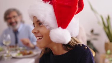 Noel şapkalı, gülümseyen beyaz kızı yakından çek. Ailenin odaklanmamış geçmişi, evdeki şenlikli yemeği kutlamak için masada toplandı. Mutlu insanlar tatil toplantısında iyi vakit geçiriyorlar..