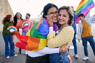 Gey gururu festivalinde birbirlerine sevgiyle sarılan lezbiyen kızların portresi. İki genç çok ırklı kadın, arka planda heyecanlı arkadaşlarıyla LGBT partisini kutlarken sevgi ve gülümseme sergiliyor..