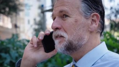 Gülümseyen orta yaşlı iş adamı ofisin dışında sessizce cep telefonuyla konuşuyor. Resmi giyimde kıdemli profesyonel açık havada cep telefonu kullanıyor. Olgun gri saçlı girişimci işten ayrıldıktan sonra mutlu..