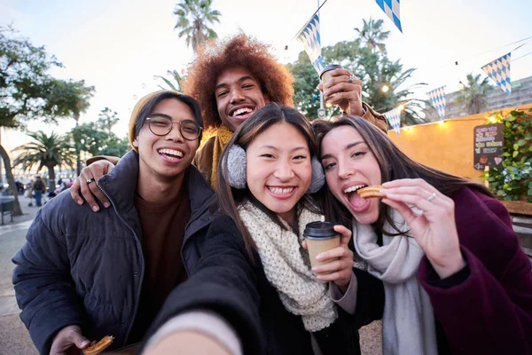 Selfie phone looking camera of cheerful friends eating chocolate churros street food stall at city outdoors. Generation z tourist group of happy people having fun in terrace cafeteria in winter time.