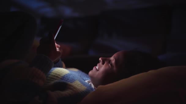 年轻的非洲女人晚上在客厅里用手机聊天 快乐的女孩沉迷于使用手机的技术 在黑暗中躺在家里 Z代的概念滚动社交媒体 — 图库视频影像