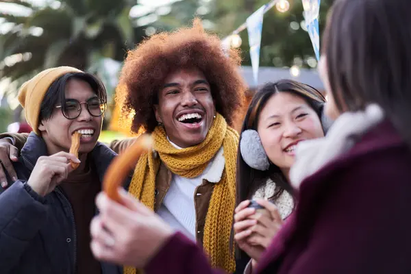 Laughing young people eating chocolate with churros together on street outdoors. Group of four multiracial friends gathered having fun at festival in winter. Generations persons holding cup hot drink.