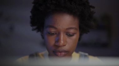 Kapalı alanda bilgisayar ekranına bakan konsantre olmuş genç afro kadının yakın çekimi. Karanlık odada dizüstü bilgisayar kullanan Afrikalı Amerikalı bir kadın. Milenyum halkı mesai saatleri dışında evden çalışıyor.. 
