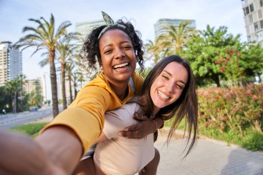 Çok ırklı lezbiyen genç çift güneşli bir günde selfie portresi çekiyorlar. Güzel, beyaz bir kadın, şehirdeki güzel Afrikalı kızı sırtında taşıyor. Gülümseyen kadın fotoğraf için mutlu poz veriyor.