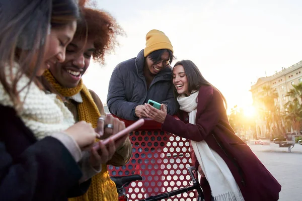 Group of young multiracial joyful people meeting cold sunny winter day chatting and using mobiles phones outdoor. Smiling happy friends enjoying amusement park. Generation z weekend activities.
