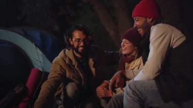 Üç gülen genç arkadaş kış kampı kaçamağında birlikte oturmaktan keyif alıyorlar. Bir grup neşeli insan kamp alanında buluşuyor. Mutlu çok ırklı milenyumlar tatil doğa gezisinde gülümsüyor..