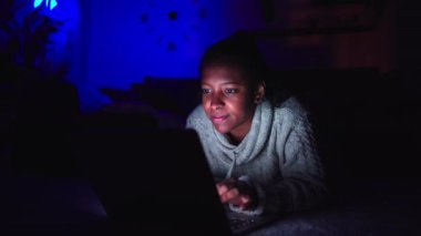 Geceleri evinde dizüstü bilgisayar kullanan neşeli, Afro-Amerikan bir kadın. Siyah kız halı kaplı oturma odasında uzanıp karanlıkta yazmaktan ve konuşmaktan mutlu oluyor. Arkaplanda mavi ışık olan insanlar.