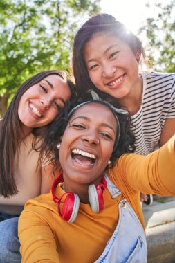 Dikey selfie üç heyecan verici çok kültürlü, neşeli genç bayan dışarıda. Kadın, gülümseyen kameraya bakıp yaz tatilinin tadını çıkarırken, lensler parlıyor. Güneşli parkta Z jenerasyonu.