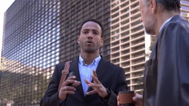 Büyük yaş farkı olan diğer uluslardan iki profesyonel erkek güneşli bir sabahta şehir caddesinde rahatça konuşuyorlar. Resmi takım elbiseli çeşitli iş adamları ofis tatilinde dışarıda çalışacaklar..