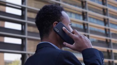 Yakışıklı Afrikalı iş adamı yürürken kameraya dönüp telefonda ciddi bir ifadeyle konuşuyor. Profesyonel takım elbiseli erkek açık havada cep telefonuyla konuşuyor. Kendine güvenen başarılı işçiyi kapat.