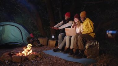 Kış gecesi kamp alanında şenlik ateşi etrafında yan yana otururken dizüstü bilgisayar kullanan üç kişi. Mutlu genç arkadaş grubu sörf ağının tadını çıkarıyor. Açık doğa kaçamağı için kamp çadırı..