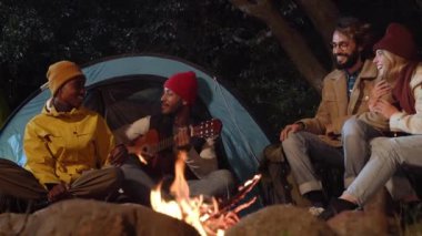 Çoklu etnik gruptan arkadaşlar kamp ateşi etrafında gülüp gitar çalıyorlar. Mutlu gençler ormanda otururken eğleniyor. Milenyum insanları vahşi doğa haftasonunda 