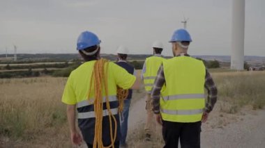 Sarı yelek ve miğfer giyen, donanımlı aletler takan, değirmenin bakımı için birlikte yürüyen mühendis işçi grubu rüzgar santralinin arka görüntüsü. Profesyonel teknisyenler, tamirden sorumlu adamlar..