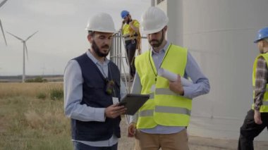 Ekip mühendisleri yel değirmeninin bakımını tartışmak için kırsal kesimdeki rüzgar santralinde tablet kullanıyorlar. Mills ve arka plandaki işçiler. Yenilenebilir enerji temiz sürdürülebilir gelecek.