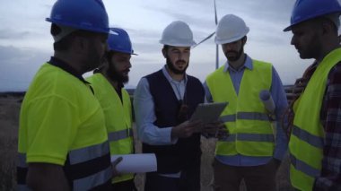 Şirket müdürü, kırsal kesimde gün batımında rüzgar santralinde tablet kullanan mühendislere açıklama yapıyor. Teknisyenler yel değirmeni bakımını güvenlik kıyafetleriyle tartışıyor. Yenilenebilir enerji temiz sürdürülebilir gelecek.