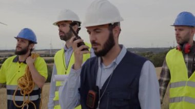 Rüzgâr santralinden bir grup mühendis güvenli giysiler giyerek ve çalışma ekipmanları taşıyarak yel değirmeninin bakımı için birlikte yürüyorlar. Onarımlardan sorumlu ciddi, konsantre adamlar..