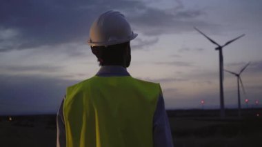 Rüzgar santralinde gün batımında dikilip türbinlerin pürüzsüz çalışmasını izleyen konsantre insan mühendisinin arka görüntüsü. Profesyonel Kafkas bakım elemanı. Güvenli kıyafetler giyiyor..