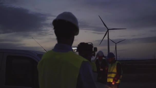 男人工程师用相机拍摄她的同事站在夕阳西下的风力发电厂看起来像磨坊 观察涡轮机的光滑 穿着安全服装的职业白人男性 — 图库视频影像