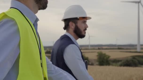 工程师组风力发电厂一起步行规划磨坊 身穿黄色背心 头戴安全帽 在风车农场配备工作工具 专业技术人员 — 图库视频影像