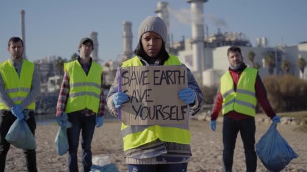 一群年轻的 多样化的示威者关注着外面的环境 严肃地看着工厂外的摄像头 黄色背心 黑人女童横幅和平抗议海滩上的污染废物 — 图库视频影像