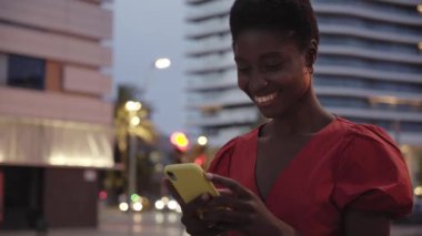 Şehir merkezinde akıllı telefon kullanan gülümseyen Afro-Afrikalı bir kadın. Sosyal ağlar yazan güzel siyah kadın. Binalar ve araba ışıkları arka plan, bağımlılık teknolojisi kavramı.