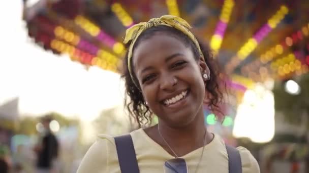 在游乐园旋转木马的背景下 年轻的非洲女孩带着友好的微笑高兴地看着相机 一个美丽的笑容可亲的女人站在外面 人们的业余时间很有趣 — 图库视频影像
