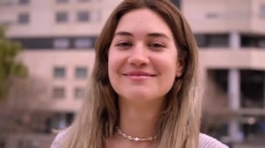 Dışarıdaki neşeli sarışın kıza yakın durun. Güçlü Avrupalı kadın gülümseyen kameraya bakarak poz veriyor. Arka planda şehir binaları olan mutlu insanlar video için poz veriyor.