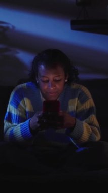 Dikey genç siyah kız gece oturma odasında akıllı telefondan sohbet ediyor. Evde karanlıkta koltukta rahat rahat oturarak teknolojiye bağımlı hale gelmiş. Sosyal medya oluşturma z kaydırılıyor.