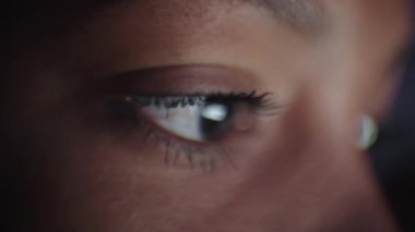 Genç siyahi kadın gözlerinin kamera ekranına bakıp bilgisayar laptopuyla çalışmasını. Monitör ışığı yüzüne yansıyor. Karanlık bir gecede, oturma odasında internette serbest çalışan bir kadın..