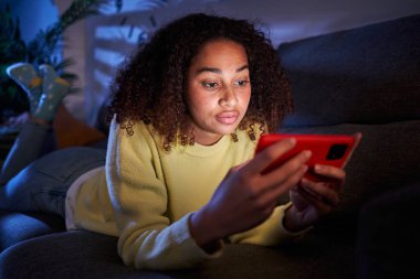 Oturma odasındaki kanepede uzanmış seyyar telefona odaklanmış genç Latin kız. Ciddi bir afro kadın gece evde cep telefonu videosu izliyor. Kadın nesli Z siyah insanlar ve yerel eğlence zamanı.