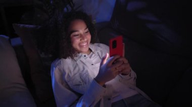 Kanepede uzanmış cep telefonu kullanan neşeli siyah kız. Hoş bir afro kadın gece evde cep telefonuyla sohbet etmekten hoşlanıyor. Bekar Afro-Amerikan kadın nesli z insanları randevu uygulamaları ve sosyal ağlarda