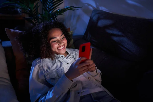 Joyful young black girl using cell phone lying on sofa. Nice afro woman enjoying chatting on mobile at night home. Single African American female generation z people on dating apps and social networks