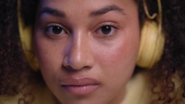 Kameraya ciddi bakan sarı kulaklıklı, sevimli, Afro-Amerikalı bir kadın. Afro kız. Düşünceli bir yüz ifadesi var. Kapalı alanda el ele poz veriyor. Latin kadınların yüzleri. 
