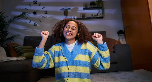 Young afro teenage girl enjoying with eyes closed favorite music in living room with headphones. Happy Latin woman dances listening to songs at home in evening. Generation z and domestic entertainment
