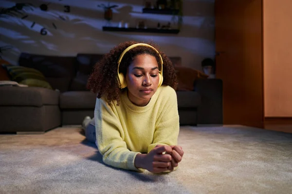 Young African American girl lying relaxed in living room listening to music yellow wireless headphones. Afro latina generation z woman calm on carpet enjoying songs at cozy home at night. Copy space